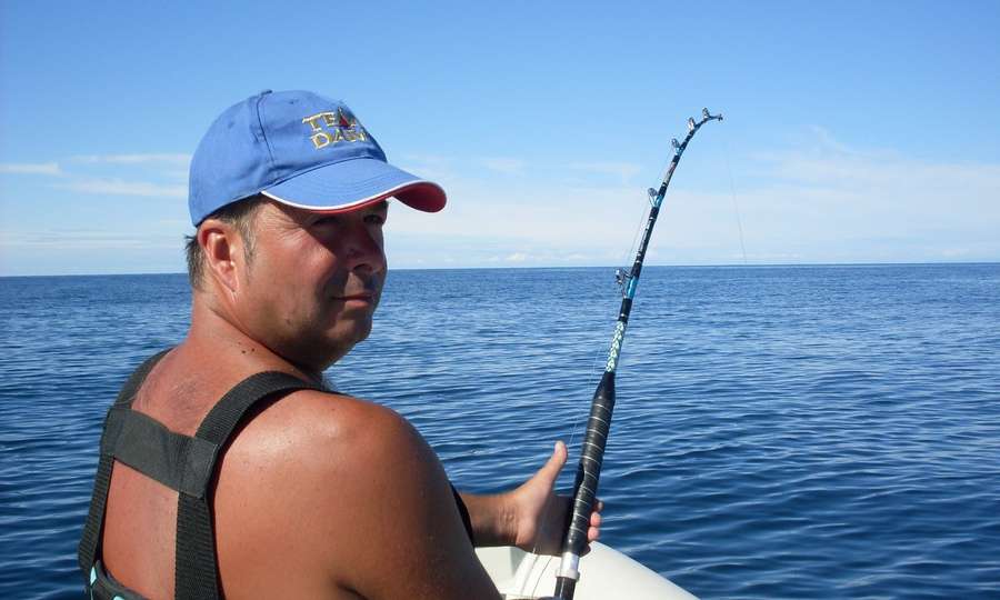 Il pescatore con l’imbragatura e la canna in curva, sul mare blu