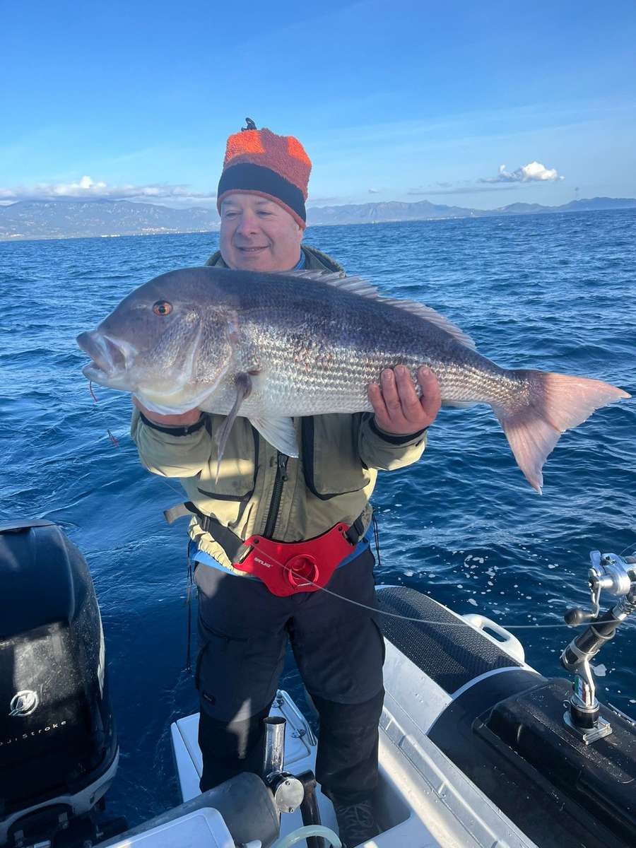 Il pescatore col berretto arancione solleva una grossa cattura, con le montagne dietro