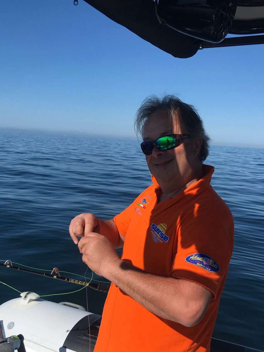 Un pescatore in maglia arancione prepara la lenza sul gommone, mare piatto