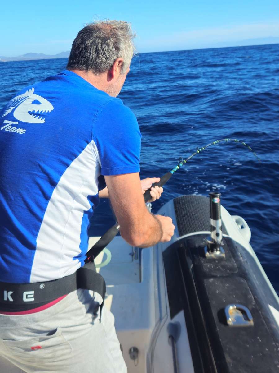 Un pescatore di spalle a poppa del gommone tiene la canna in curva, mare blu e costa in lontananza