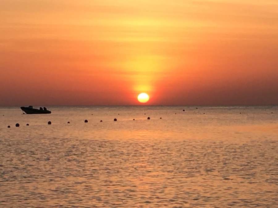 Il sole basso sull’orizzonte, cielo arancio, un gommone in controluce e una fila di boe